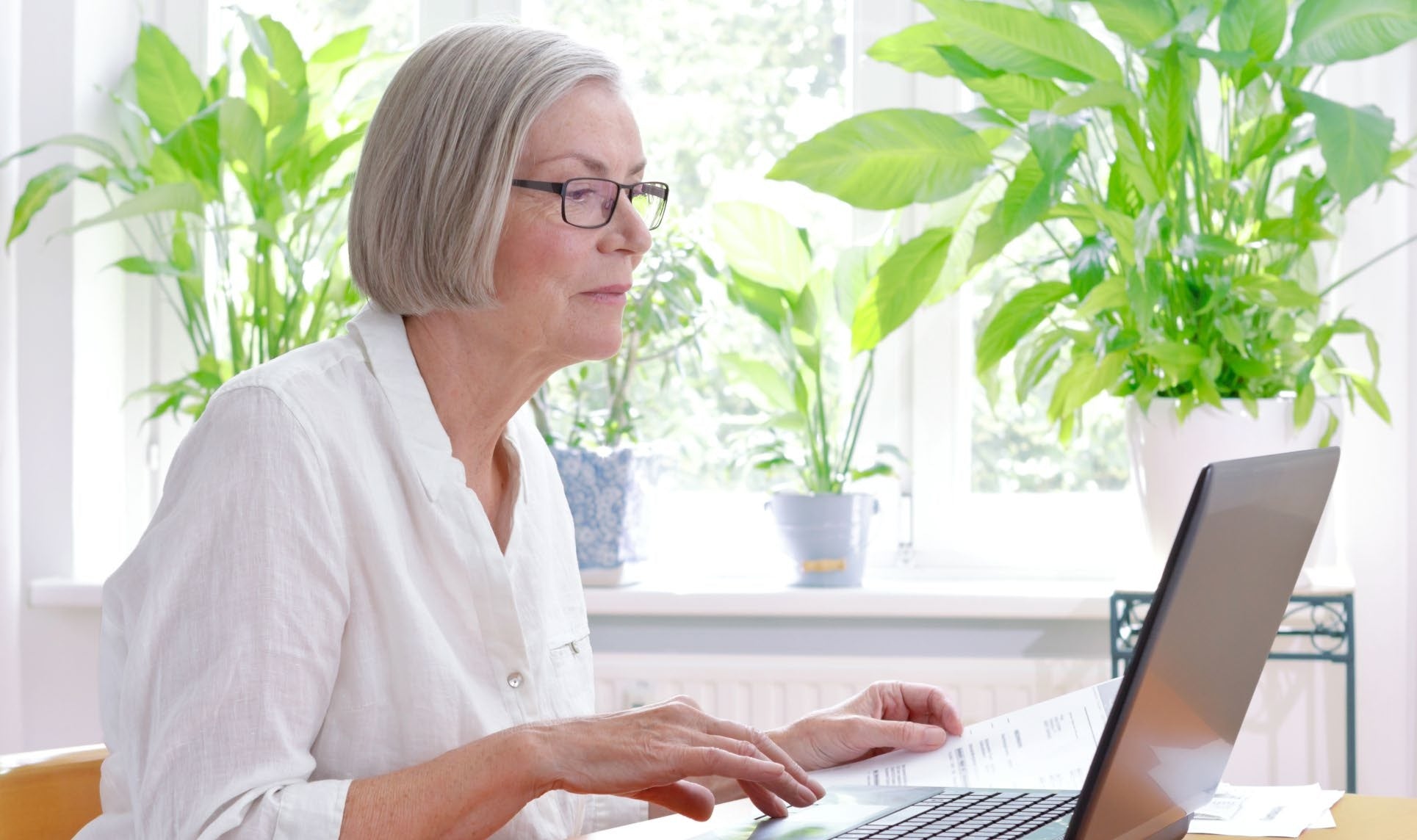 An older woman wearing glasses while using a laptop beside a bright window